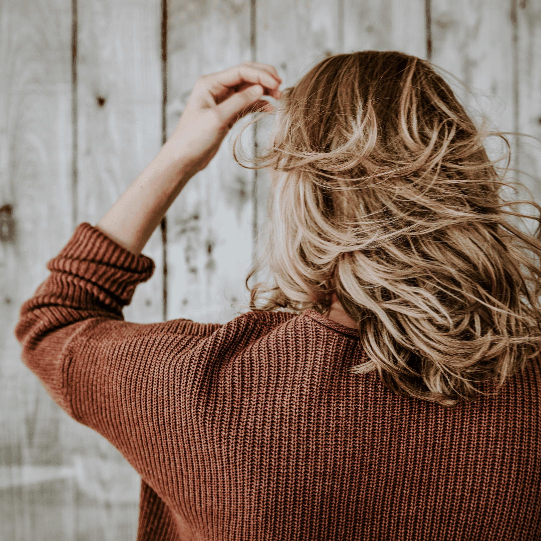 Woman with wavy blonde hair in a brown sweater, facing away, against a wooden backdrop.
