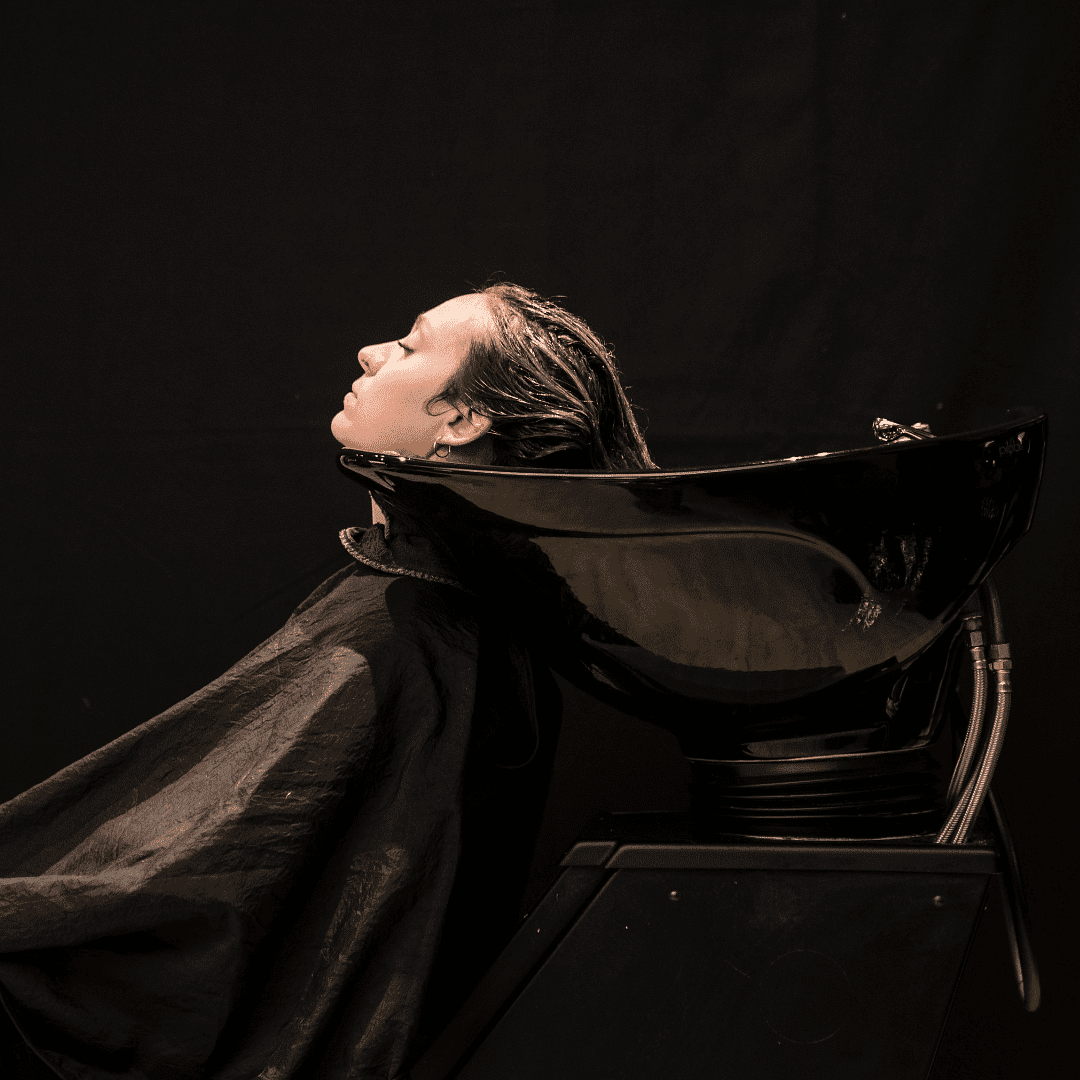 Person with wet hair reclining at a salon sink under a black cape, receiving a hair wash.