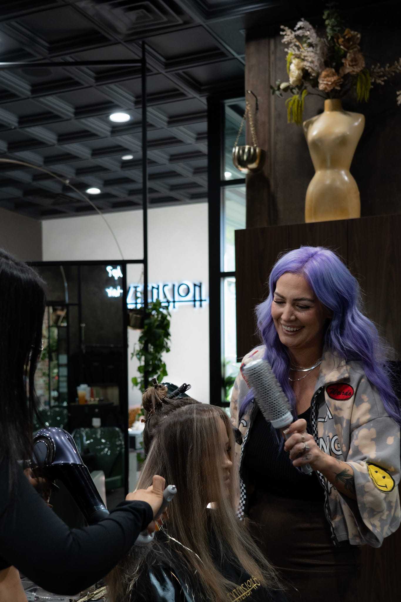 Hair salon scene with stylist blow-drying client's hair, another stylist holds hairbrush.