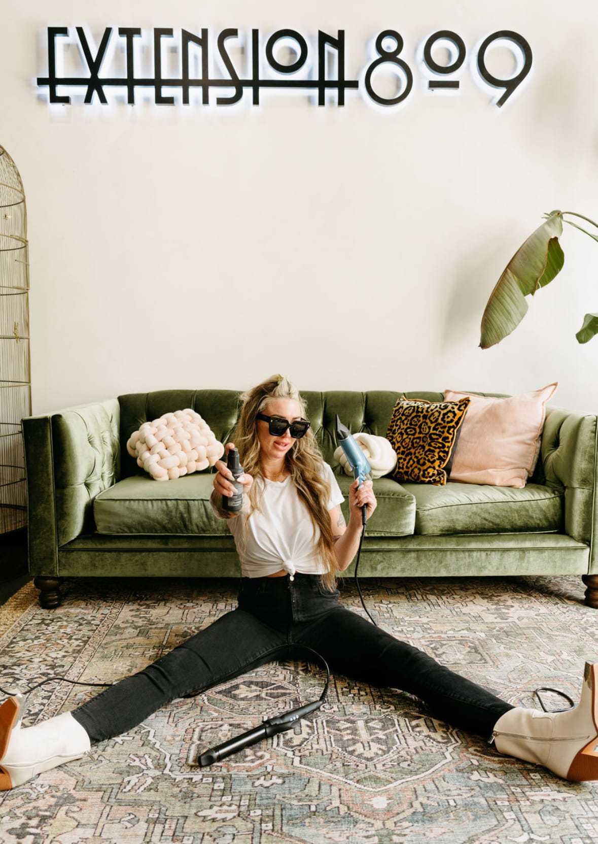Woman sitting on floor, styling hair with tools, in front of a green sofa.