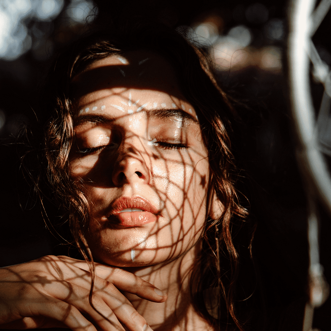 Woman with eyes closed, sunlight filtering through patterned shadows on her face.