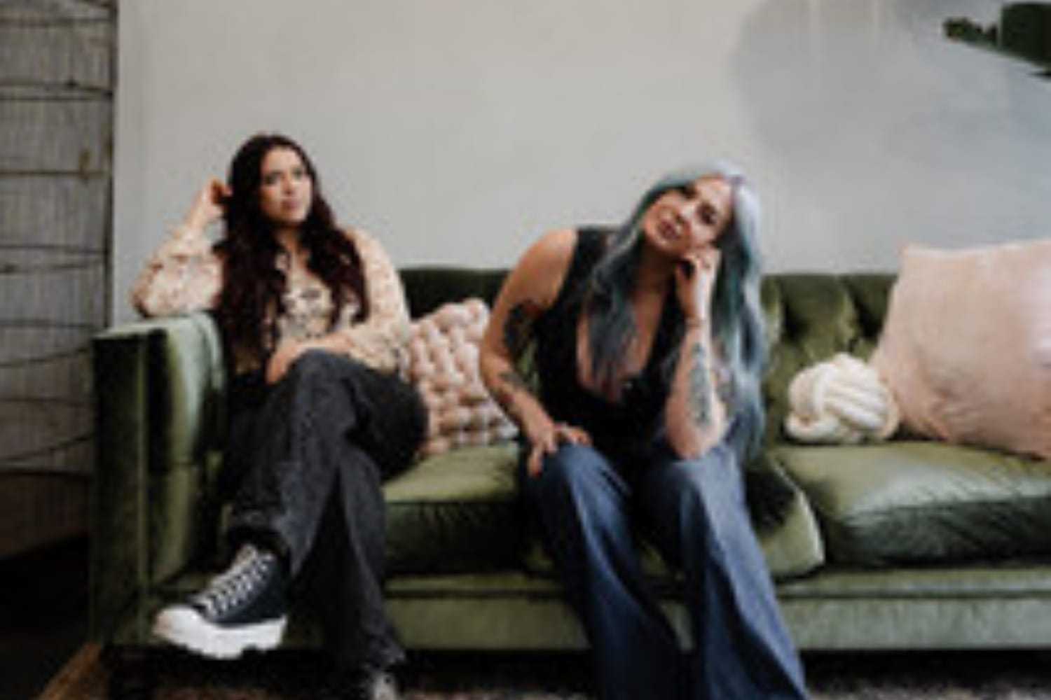 Two women sitting on a green sofa, one with tattoos, both casually posed.