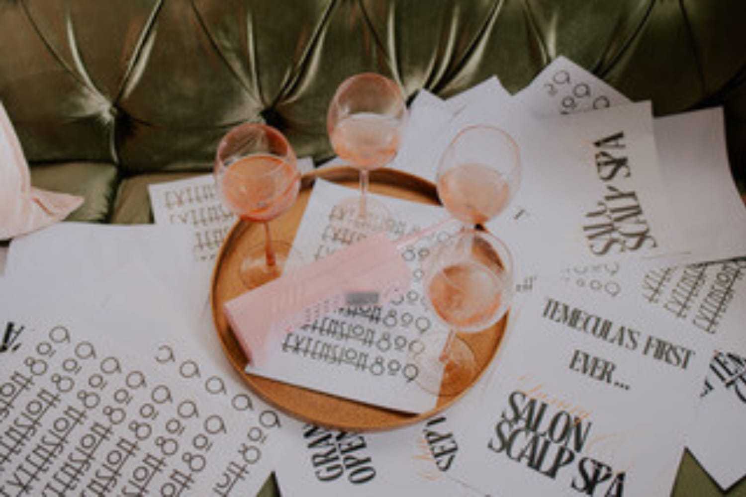 Pink wine glasses on a tray with scattered design papers on a velvet green sofa.