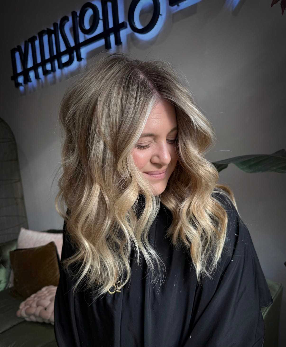 Blonde woman with wavy hair sitting in a salon chair, smiling contentedly.